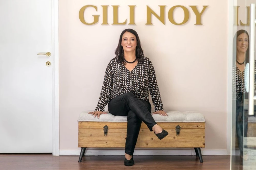 אישה יושבת על ספסל מרופד מתחת לשלט "GILI NOY", מחייכת, בחדר מודרני ומואר היטב.