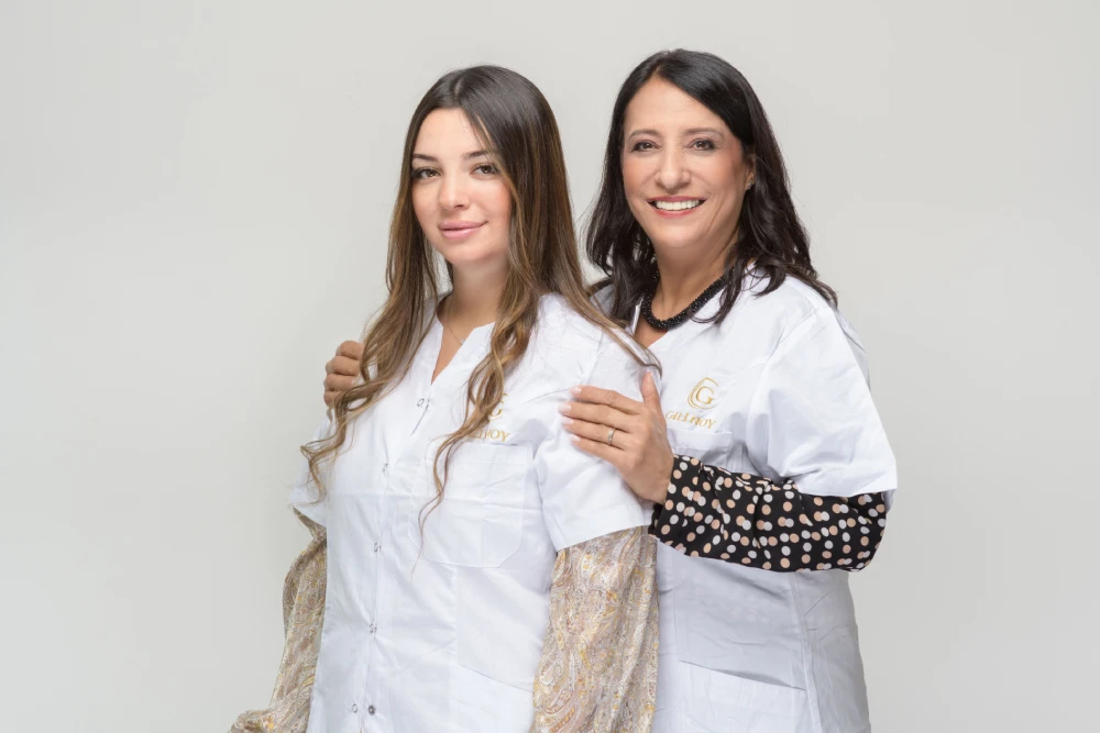 שתי נשים לבושות בחלוקים לבנים, מחייכות ועומדות זו לצד זו על רקע בהיר ופשוט.