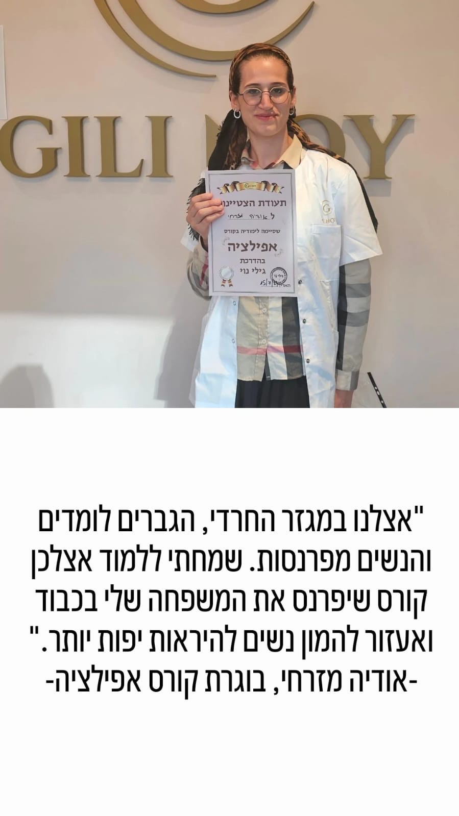 אישה במעיל לבן מחזיקה תעודה ומחייכת, בעודה עומדת מול השלט "GILI ALOY".