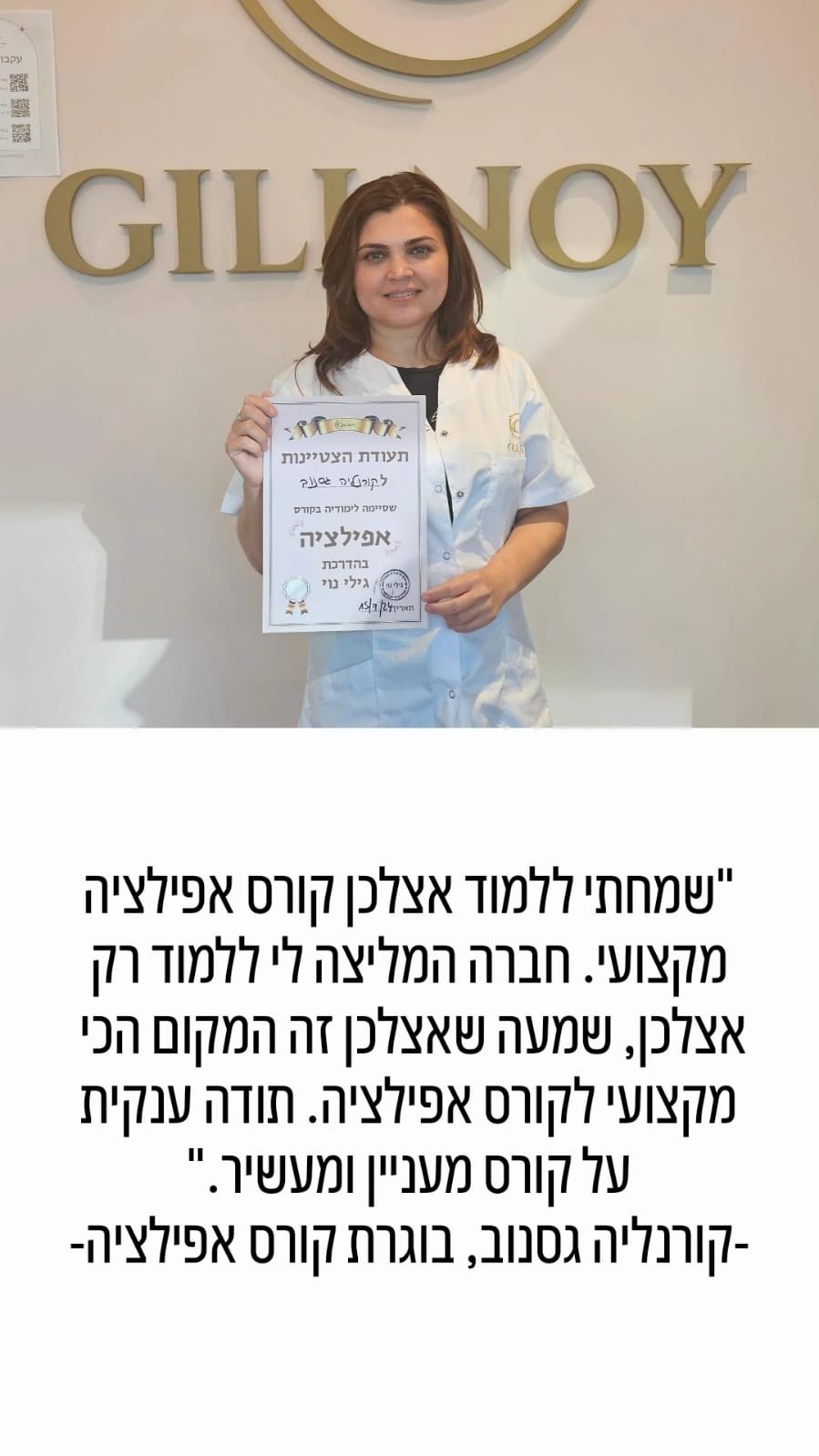 אישה לבושה במעיל לבן מחזיקה תעודה ומחייכת, בעודה עומדת מול שלט "GILINOY" התלוי על הקיר.