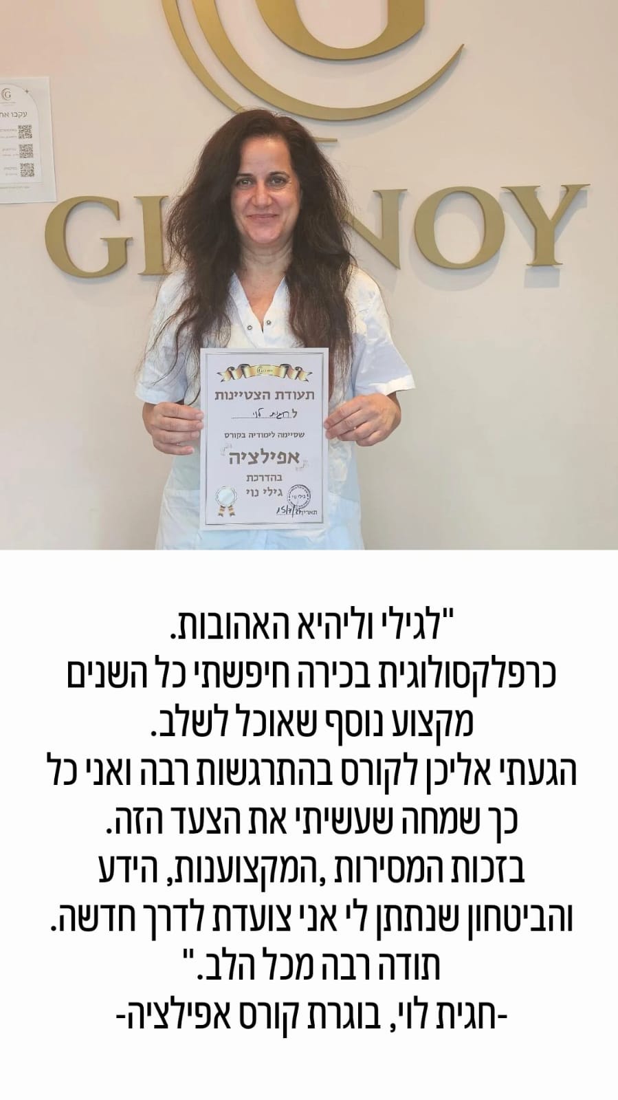 אישה עם שיער ארוך וכהה, מחזיקה תעודה, מחייכת, עומדת מול קיר שעליו כתוב טקסט.
