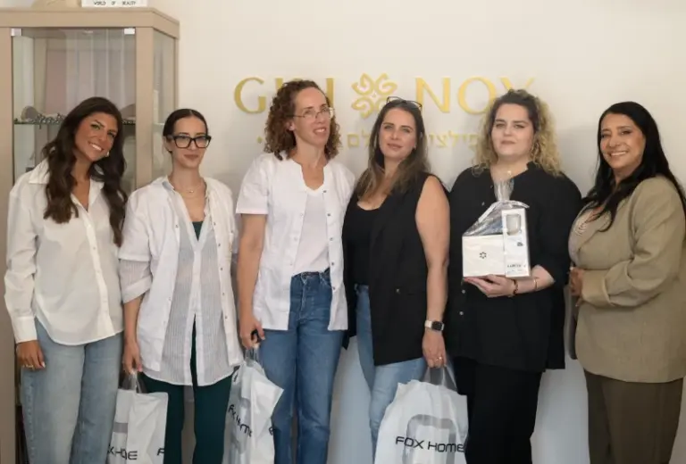 שש נשים עומדות יחד בתוך מבנה, אוחזות בשקיות מתנה, ומחייכות מול קיר שעליו מוטבע לוגו זהוב.