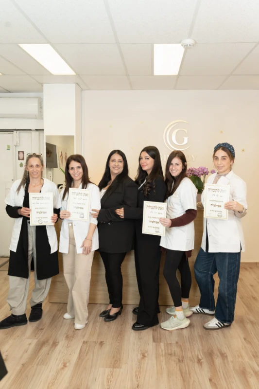 שש נשים עומדות בתוך הבית, ארבע מהן מחזיקות תעודות, וכולן מחייכות למצלמה בחדר מואר עם רצפת עץ.