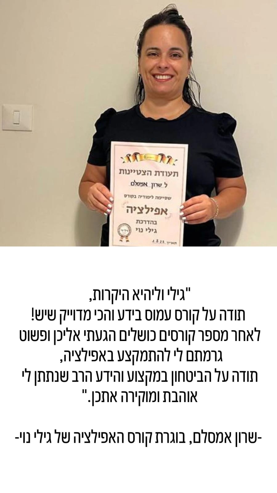 אישה מחייכת כשהיא אוחזת בתעודה, עומדת על רקע קיר חלק, לבושה בחולצה כהה.
