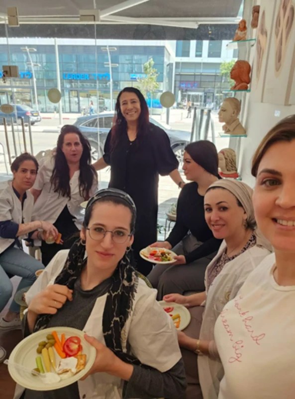 שבע נשים, חלקן לבושות בחלוקים לבנים, יושבות בחדר מואר ומודרני, חולקות ארוחה ומחייכות למצלמה.