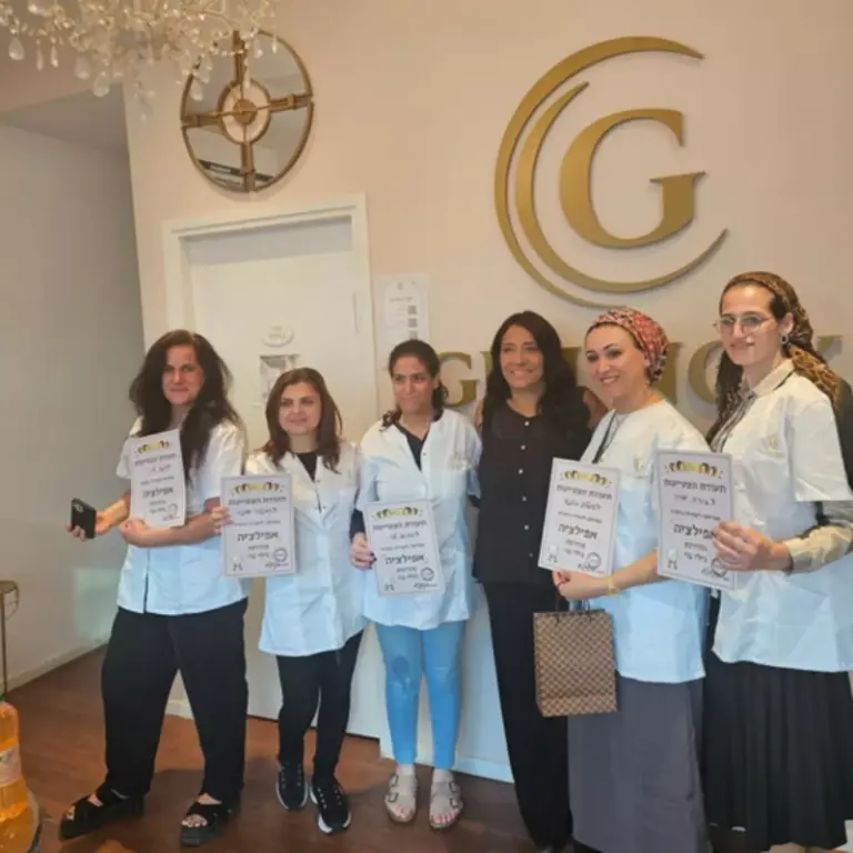 שש נשים עומדות יחד בתוך חדר, חמש מהן מחזיקות תעודות, וכולן מחייכות מול לוגו התלוי על הקיר.